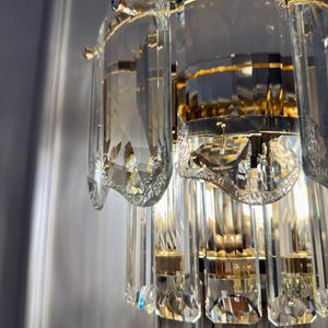 May include: Close-up of a crystal wall sconce with a gold-colored metal frame. The sconce features multiple tiers of rectangular and curved crystal elements, reflecting light. The design is ornate and elegant, suitable for interior decor.