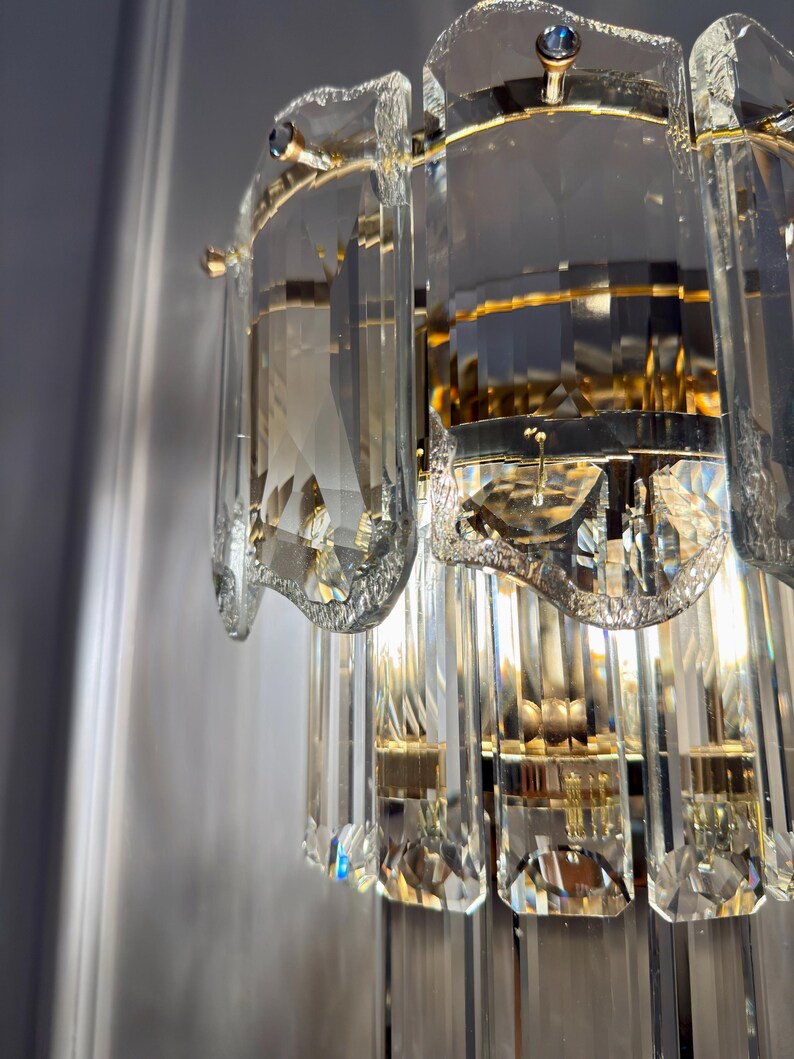 May include: Close-up of a crystal wall sconce with a gold-colored metal frame. The sconce features multiple tiers of rectangular and curved crystal elements, reflecting light. The design is ornate and elegant, suitable for interior decor.