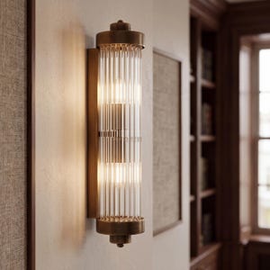 May include: A cylindrical wall sconce featuring a vertical ribbed glass shade encased in a brushed bronze metal frame. The fixture has a flat rectangular backplate and rounded top and bottom caps, emitting a warm glow from internal light bulbs.