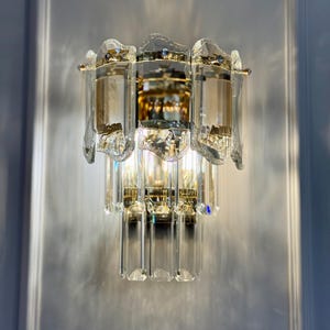 May include: A crystal and gold wall sconce with a multi-tiered design. The sconce features clear, faceted crystal elements and gold-toned metal accents. The light fixture is mounted on a light gray wall, creating a luxurious aesthetic.