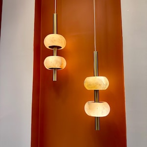 May include: Two pendant lights with a modern design. Each light features three stacked, oval-shaped, cream-colored shades. The shades are connected by a brass-colored rod and suspended from a white cord. The background is a warm, reddish-brown color.