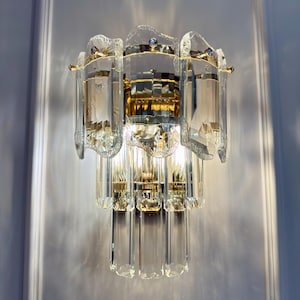 May include: A crystal wall sconce with a gold-toned metal frame. The sconce features multiple clear, rectangular crystal elements and a decorative, layered design. The light fixture is mounted on a white wall.