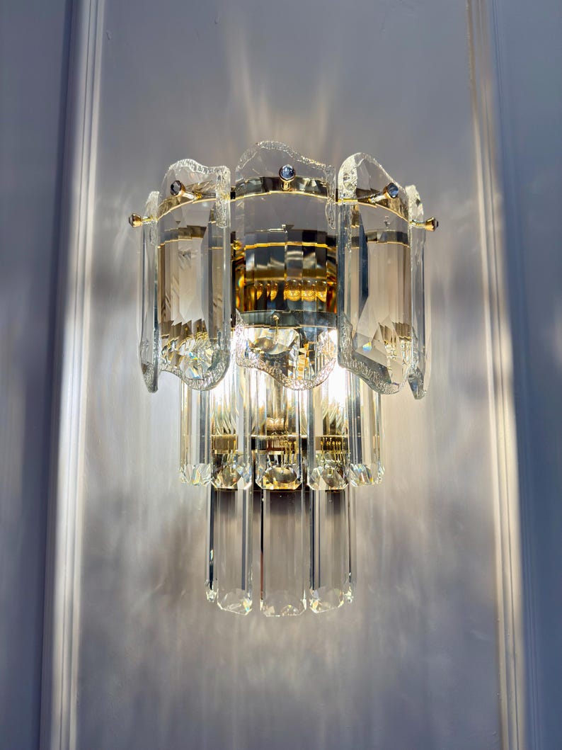 May include: A crystal wall sconce with a gold-toned metal frame. The sconce features multiple clear, rectangular crystal elements and a decorative, layered design. The light fixture is mounted on a white wall.