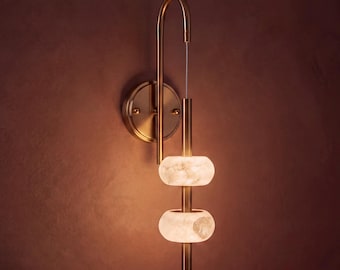 May include: A wall sconce featuring a curved bronze-toned metal arm and a circular mounting plate. Two stacked, glowing, translucent white stone rings are suspended along a vertical metal rod, emitting a soft, warm light from within the stone.