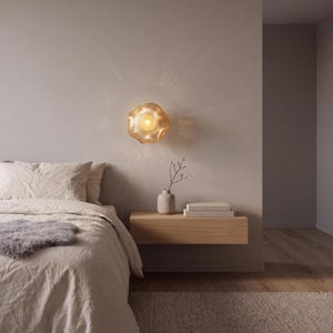 May include: A wall-mounted light fixture with a translucent, wavy, amber-toned glass shade that emits a warm, glowing light, creating a soft, radiating pattern on the surrounding wall surface above a wooden floating nightstand.