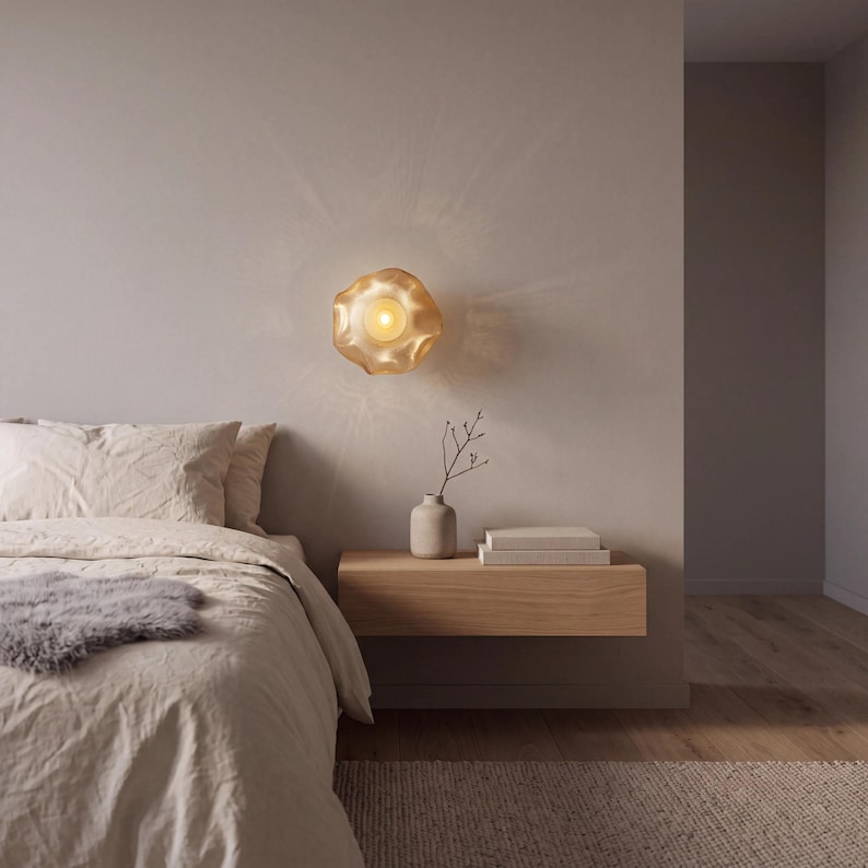 May include: A wall-mounted light fixture with a translucent, wavy, amber-toned glass shade that emits a warm, glowing light, creating a soft, radiating pattern on the surrounding wall surface above a wooden floating nightstand.