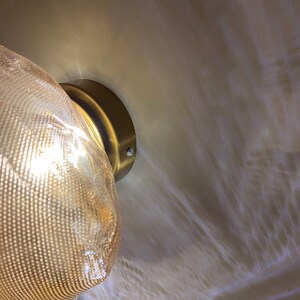 May include: A wall-mounted light fixture featuring a textured, translucent amber-toned glass shade with a fine mesh pattern. The shade is attached to a circular, brushed brass-colored metal base, casting a warm, diffused glow.