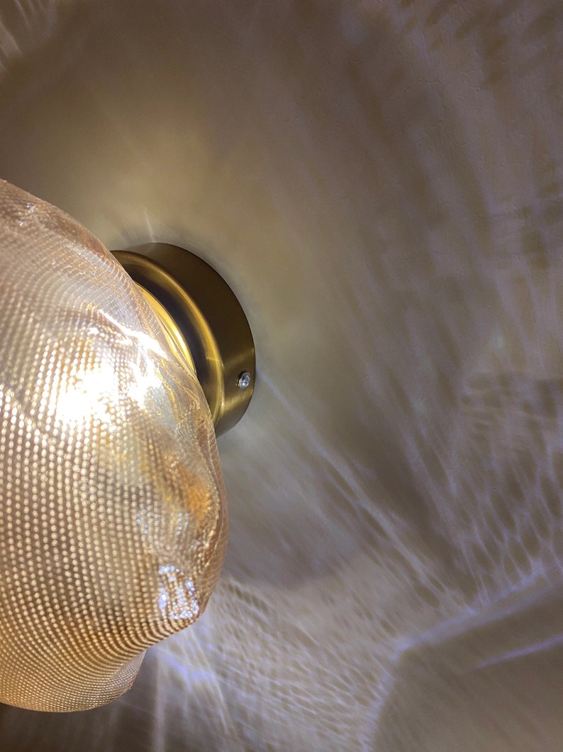 May include: A wall-mounted light fixture featuring a textured, translucent amber-toned glass shade with a fine mesh pattern. The shade is attached to a circular, brushed brass-colored metal base, casting a warm, diffused glow.