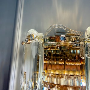 May include: Close-up of a crystal and gold-toned wall sconce. The sconce features multiple clear, faceted crystal elements and gold-colored metal accents. The design includes a curved top and a rectangular base, reflecting light.