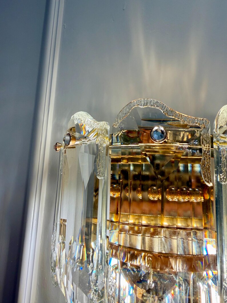 May include: Close-up of a crystal and gold-toned wall sconce. The sconce features multiple clear, faceted crystal elements and gold-colored metal accents. The design includes a curved top and a rectangular base, reflecting light.