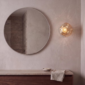 May include: A large circular wall mirror with a thin metal frame positioned next to an organic-shaped, amber-tinted glass wall sconce that emits a warm, glowing light, creating a soft radiance on the textured, light-colored wall surface.