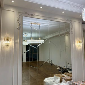 May include: A large, rectangular mirror composed of multiple vertical panels, installed on a white wall with decorative molding. The mirror reflects a room with a hardwood floor and a chandelier. Two sconces flank the mirror.