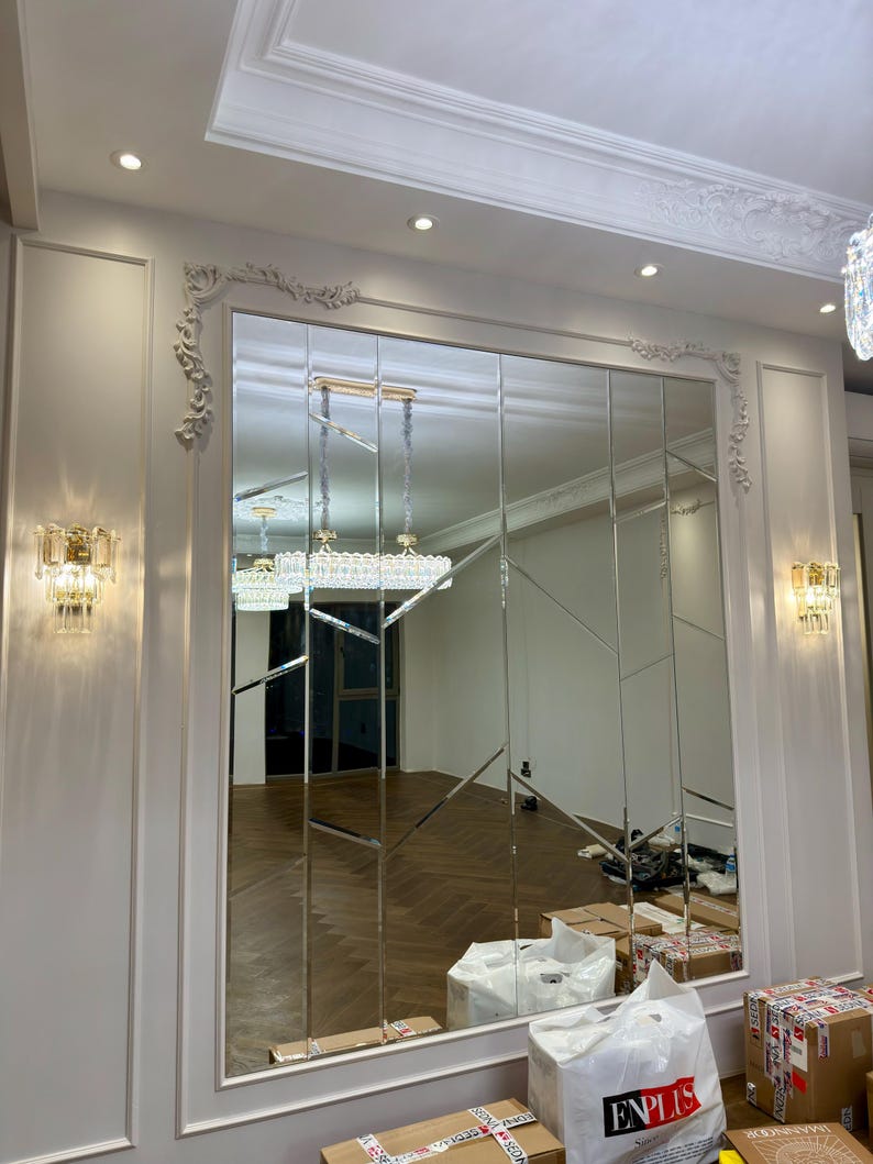 May include: A large, rectangular mirror composed of multiple vertical panels, installed on a white wall with decorative molding. The mirror reflects a room with a hardwood floor and a chandelier. Two sconces flank the mirror.
