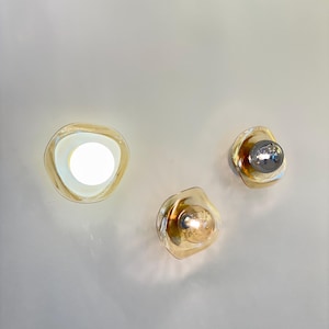 K&ouml;nnte beinhalten: Drei Wandleuchten mit unregelm&auml;&szlig;ig geformten, bernsteinfarbenen Glasschirmen. Eine Leuchte strahlt hellwei&szlig;es Licht aus, w&auml;hrend die anderen beiden sichtbare Leuchtmittel mit metallischen und warmen Oberfl&auml;chen enthalten.