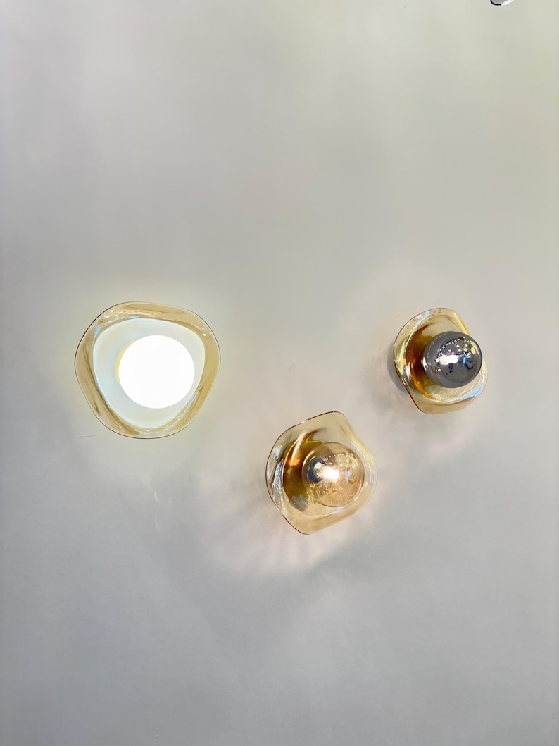 K&ouml;nnte beinhalten: Drei Wandleuchten mit unregelm&auml;&szlig;ig geformten, bernsteinfarbenen Glasschirmen. Eine Leuchte strahlt hellwei&szlig;es Licht aus, w&auml;hrend die anderen beiden sichtbare Leuchtmittel mit metallischen und warmen Oberfl&auml;chen enthalten.