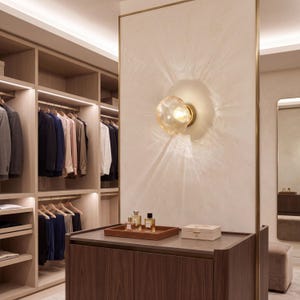 May include: A wall-mounted light fixture with a spherical, textured amber glass shade that emits a warm glow, casting radial light patterns onto a light-colored wall panel above a dark wood cabinet with perfume bottles.