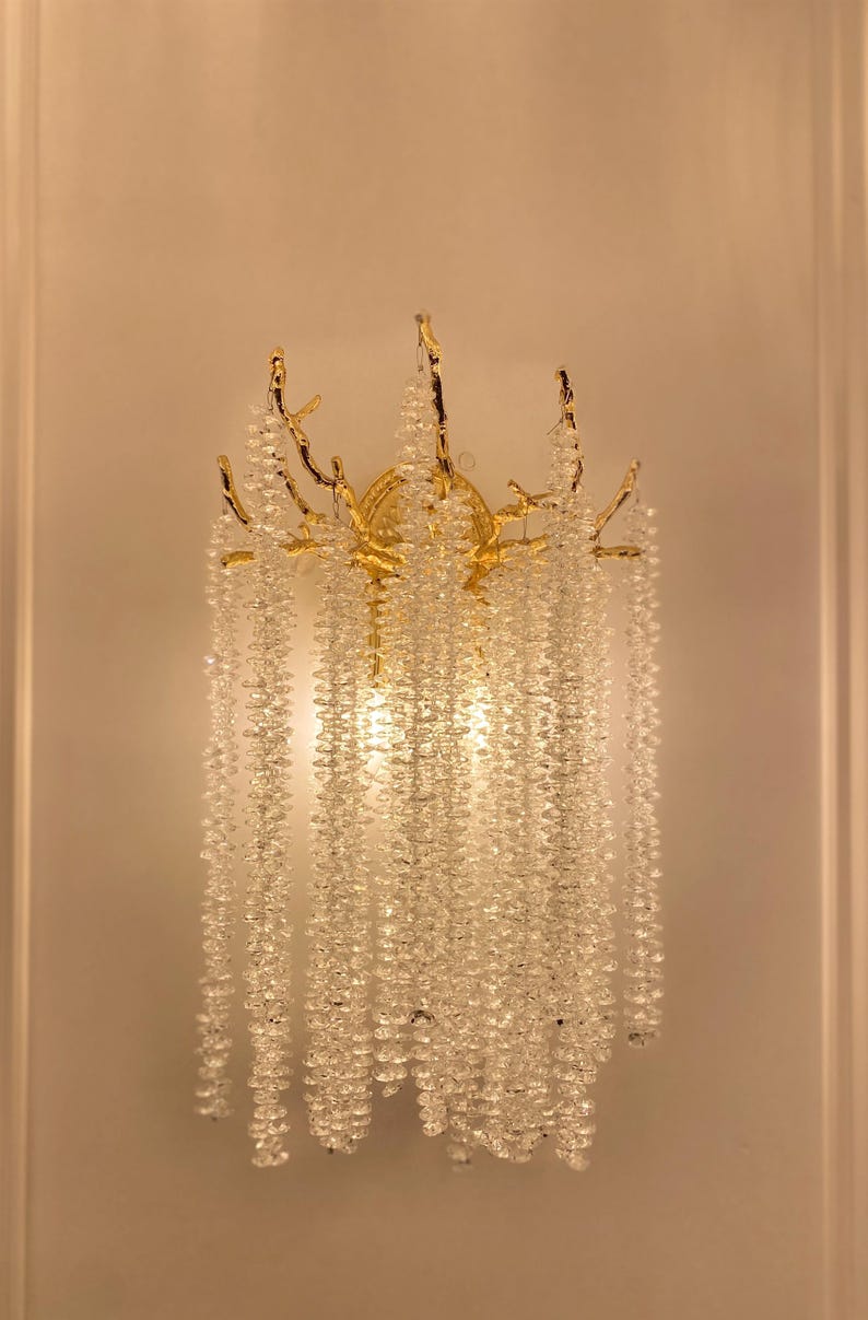 May include: A gold-toned wall sconce with a crystal chandelier design. The sconce features a branch-like structure with cascading crystal beads.