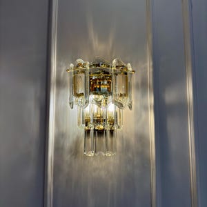 May include: A wall-mounted light fixture with a tiered design. The fixture features multiple clear, rectangular glass panels and gold-colored metal accents. The light casts a pattern on the wall. The overall style is ornate and decorative.