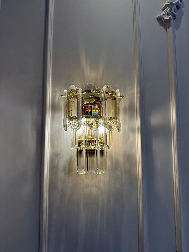 May include: A wall-mounted light fixture with a tiered design. The fixture features multiple clear, rectangular glass panels and gold-colored metal accents. The light casts a pattern on the wall. The overall style is ornate and decorative.
