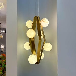 May include: A modern gold-toned chandelier with a diamond-shaped frame. The fixture features multiple spherical, glowing lights, some with a marbled appearance, suspended from thin wires. The design is contemporary, suitable for interior lighting.
