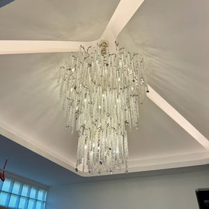 May include: A large, ornate chandelier with multiple tiers of clear, sparkling crystals hangs from a white ceiling. The chandelier's design features cascading strands of crystals, creating a dazzling display of light and reflection. The fixture is a statement piece.