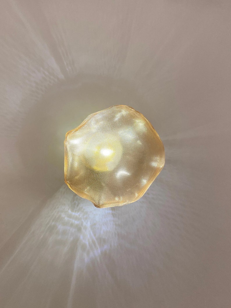 May include: A circular pendant light fixture featuring a fine gold-toned metal mesh shade. The light source within emits a warm glow, casting intricate, radiating patterns of light and shadow onto the surrounding area from the mesh texture.