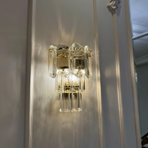 May include: A two-tiered wall sconce with a gold-toned metal frame and clear, faceted glass panels. The sconce is mounted on a white wall with molding. The light is on, casting shadows.
