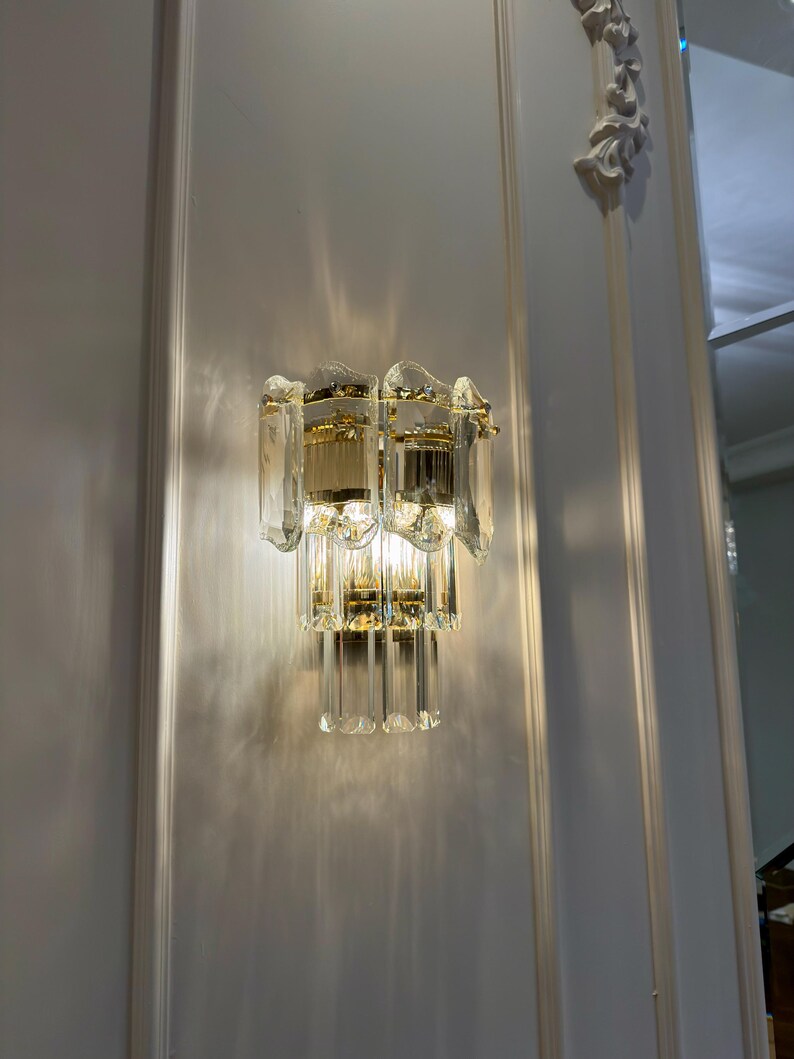 May include: A two-tiered wall sconce with a gold-toned metal frame and clear, faceted glass panels. The sconce is mounted on a white wall with molding. The light is on, casting shadows.