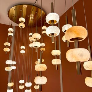 May include: A modern chandelier with multiple light fixtures. The lights have a layered, oval design with a warm, off-white glow. The fixtures are suspended from a gold-colored ceiling plate and hang at varying lengths, creating a cascading effect.