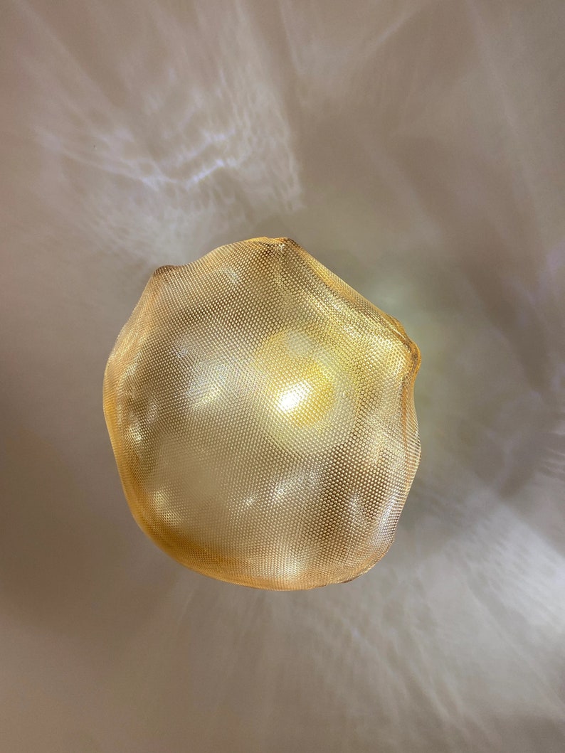 May include: A glowing, spherical pendant light fixture featuring a fine, gold-toned metallic mesh exterior. The light emits a warm, diffused radiance, casting soft, intricate patterns of light and shadow onto the surrounding area.