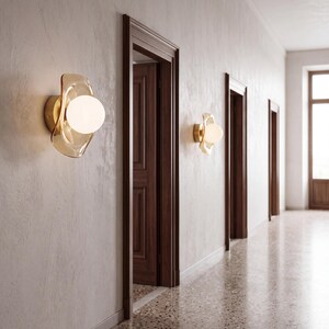 K&ouml;nnte beinhalten: Zwei Wandleuchten an einer hellen Wand. Jede Leuchte besteht aus einer runden, opaken wei&szlig;en Glaskugel, die in einer durchscheinenden, bernsteinfarbenen Glasscheibe mit organischer Form sitzt und an einem goldfarbenen Metallsockel befestigt ist.