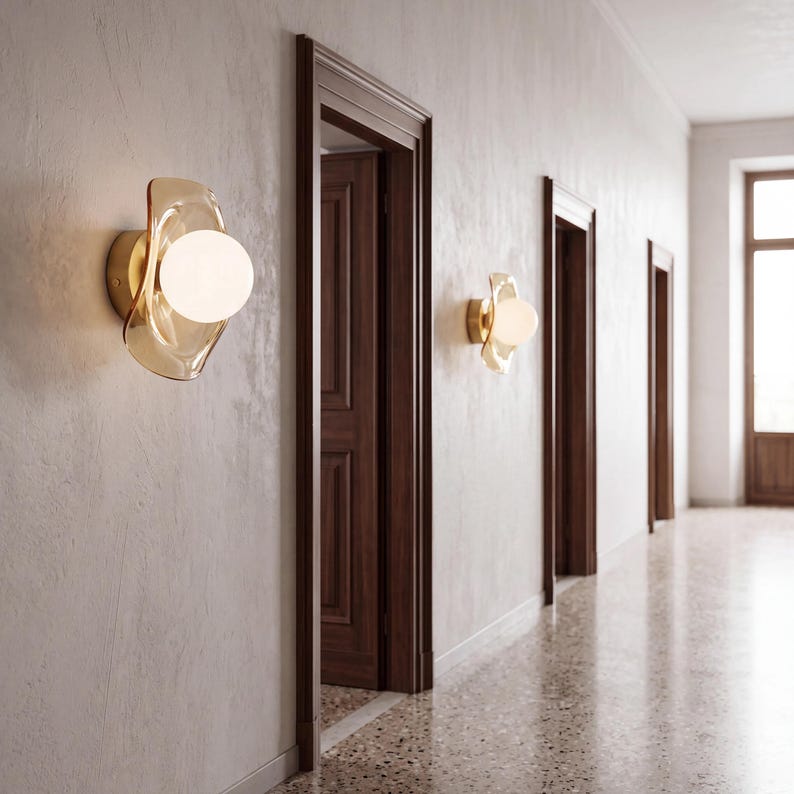 K&ouml;nnte beinhalten: Zwei Wandleuchten an einer hellen Wand. Jede Leuchte besteht aus einer runden, opaken wei&szlig;en Glaskugel, die in einer durchscheinenden, bernsteinfarbenen Glasscheibe mit organischer Form sitzt und an einem goldfarbenen Metallsockel befestigt ist.