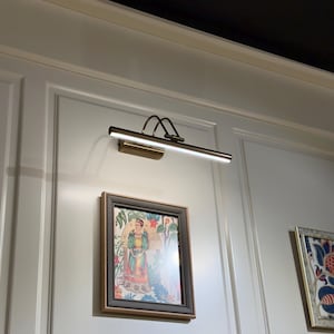 May include: A wall-mounted picture light illuminates a framed artwork. The light fixture has a rectangular head and a curved arm, finished in a brass tone. The artwork features a colorful illustration of a person in a garden setting.