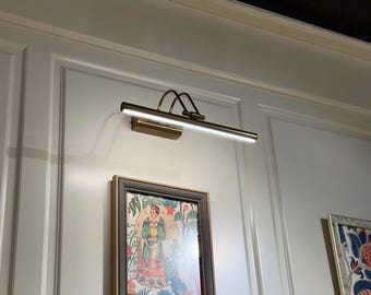May include: A wall-mounted picture light illuminates a framed artwork. The light fixture has a rectangular head and a curved arm, finished in a brass tone. The artwork features a colorful illustration of a person in a garden setting.