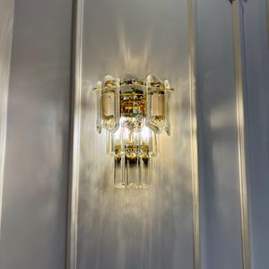 May include: A wall-mounted light fixture with a tiered design. The fixture features clear and amber-colored glass crystals and gold-toned metal accents. The light casts a pattern on the white wall.