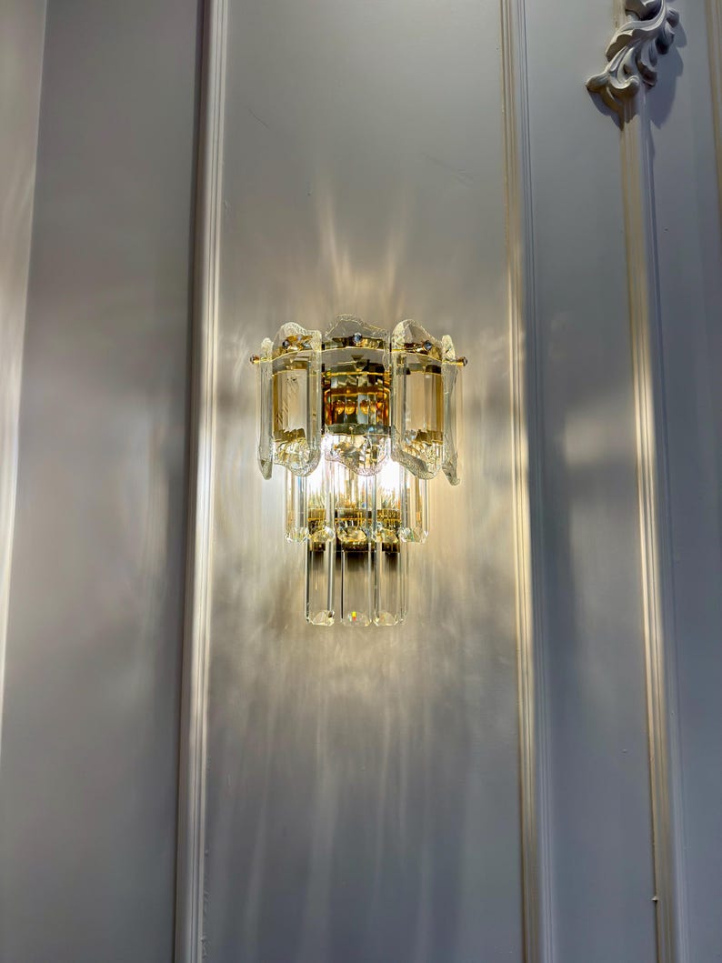 May include: A wall-mounted light fixture with a tiered design. The fixture features clear and amber-colored glass crystals and gold-toned metal accents. The light casts a pattern on the white wall.