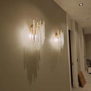 May include: Two crystal and gold wall sconces with cascading crystal beads, mounted on a light beige wall in a hallway.
