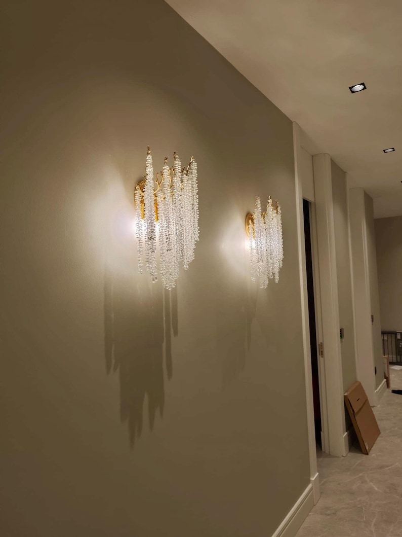 May include: Two crystal and gold wall sconces with cascading crystal beads, mounted on a light beige wall in a hallway.