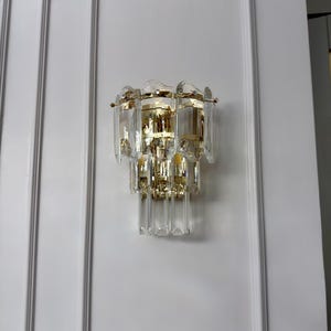 May include: A wall-mounted light fixture with a gold-colored metal frame and multiple clear, rectangular crystal accents. The fixture is mounted on a white wall with decorative molding.