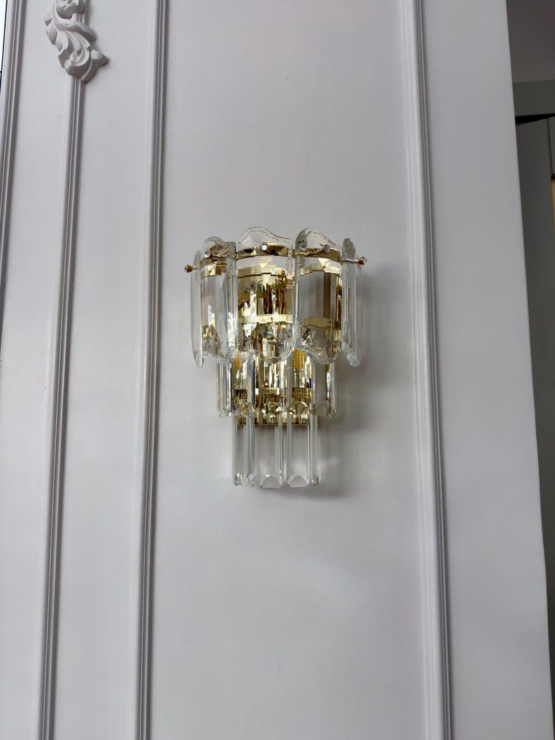 May include: A wall-mounted light fixture with a gold-colored metal frame and multiple clear, rectangular crystal accents. The fixture is mounted on a white wall with decorative molding.