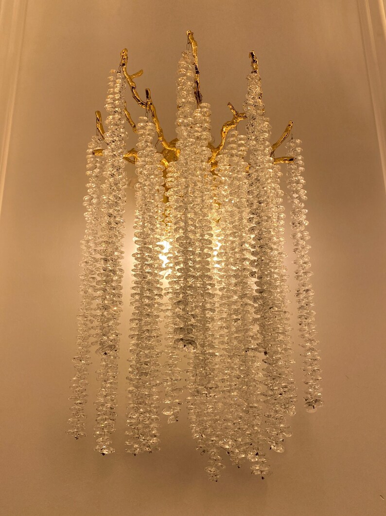 May include: A gold wall sconce with a tree branch design and crystal beads hanging down. The sconce is lit and the beads are reflecting the light.