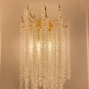 May include: A gold-toned wall sconce with a branch-like design and cascading crystal beads. The sconce is lit and the crystals sparkle.