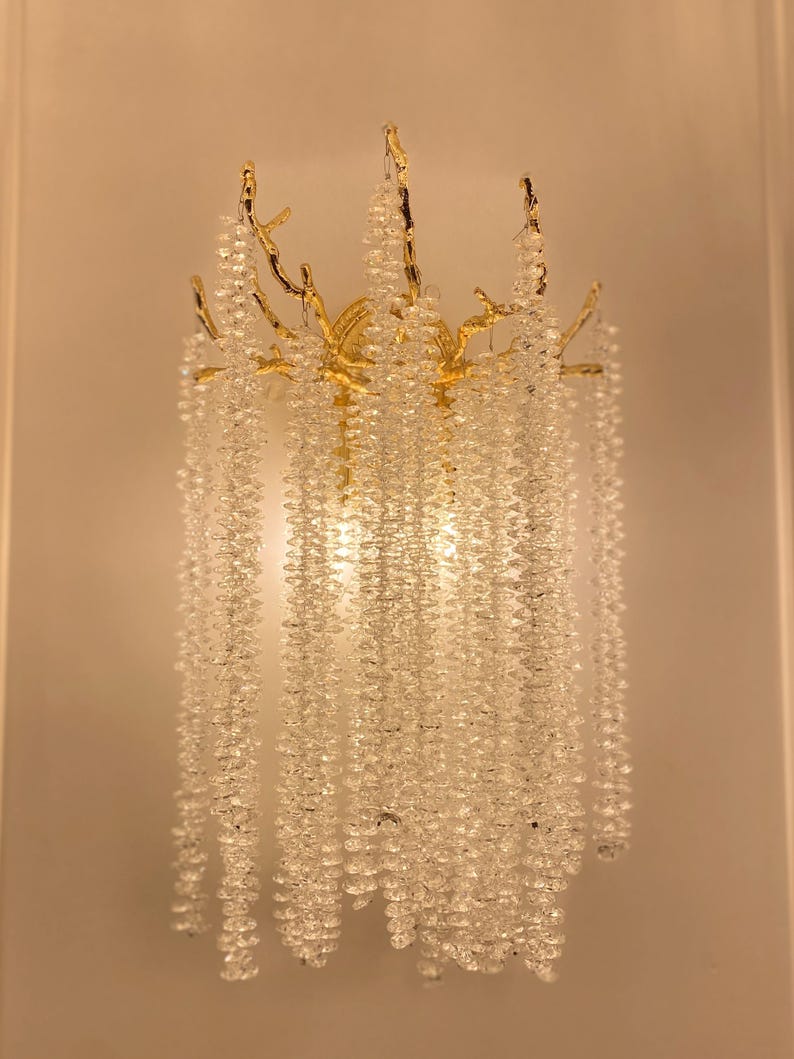 May include: A gold-toned wall sconce with a branch-like design and cascading crystal beads. The sconce is lit and the crystals sparkle.