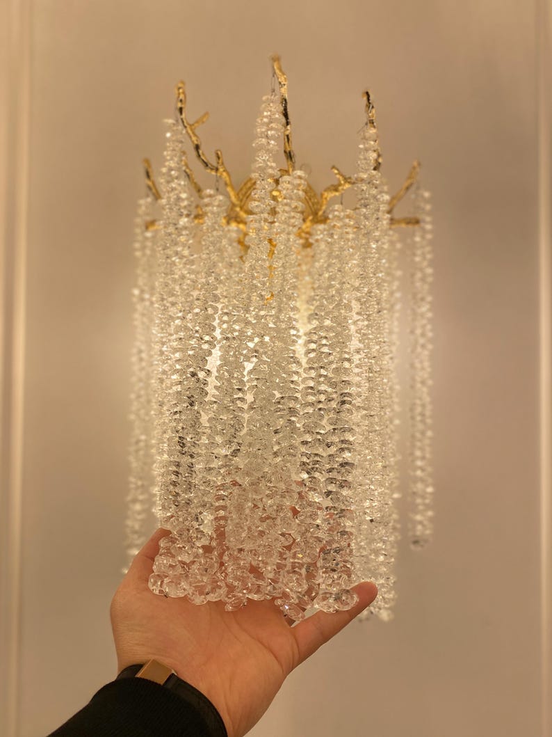 May include: A gold-toned metal chandelier with clear crystal beads hanging from the branches. The chandelier is designed to look like a tree with branches and leaves.