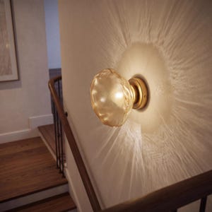 May include: A wall-mounted light fixture featuring a round, textured, amber-tinted glass shade attached to a circular brass-colored base. The illuminated glass emits a warm glow, casting a distinct, radiating light pattern onto the wall.