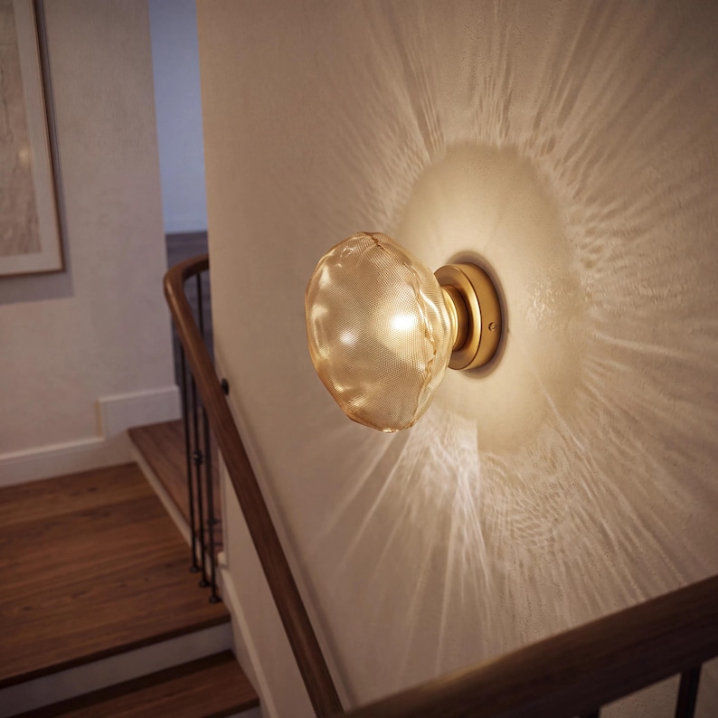 May include: A wall-mounted light fixture featuring a round, textured, amber-tinted glass shade attached to a circular brass-colored base. The illuminated glass emits a warm glow, casting a distinct, radiating light pattern onto the wall.