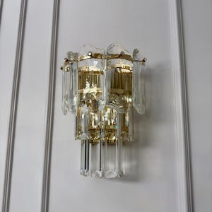 May include: A vintage wall sconce with a gold-toned metal frame and clear crystal accents. The sconce has multiple tiers of rectangular and teardrop-shaped crystals, creating a cascading effect. The fixture is mounted on a white wall.