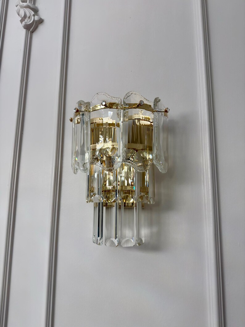 May include: A vintage wall sconce with a gold-toned metal frame and clear crystal accents. The sconce has multiple tiers of rectangular and teardrop-shaped crystals, creating a cascading effect. The fixture is mounted on a white wall.