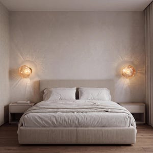 May include: A bedroom featuring a beige upholstered bed with white bedding, flanked by two matching bedside tables. Two amber-colored, organic-shaped glass wall sconces are mounted on the wall, casting a warm, radiant light pattern.