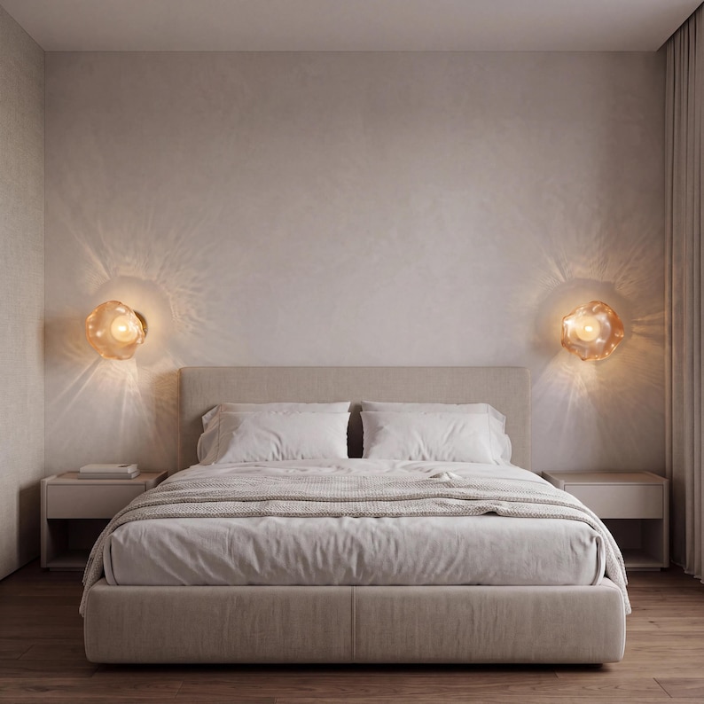 May include: A bedroom featuring a beige upholstered bed with white bedding, flanked by two matching bedside tables. Two amber-colored, organic-shaped glass wall sconces are mounted on the wall, casting a warm, radiant light pattern.